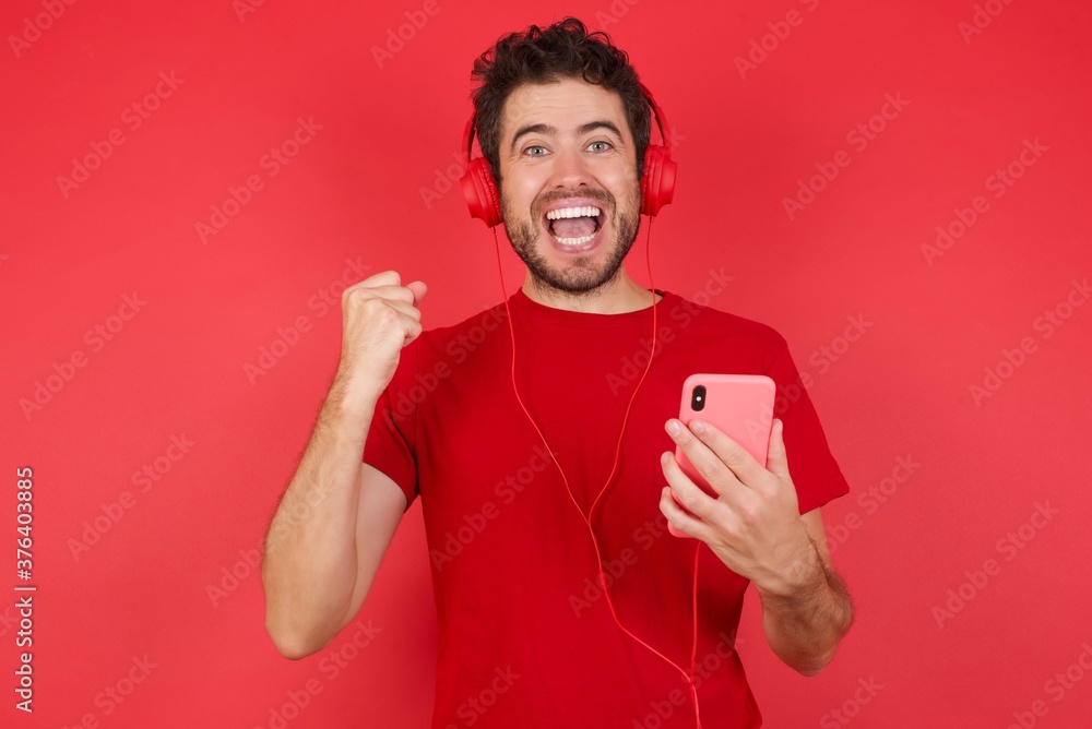 Positive Young handsome caucasian man wearing t-shirt over isolated red background holds modern cell phone connected to headphones, clenches fist from good emotions, exclaims with joy, Positive Young handsome caucasian man wearing t-shirt over isolated red background holds modern cell phone connected to headphones, clenches fist from good emotions, exclaims with joy,