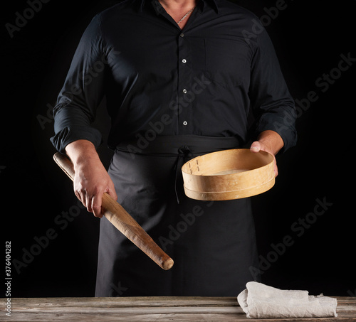 man in black uniform holding empty vintage round wooden sieve for sifting flour and rolling pin