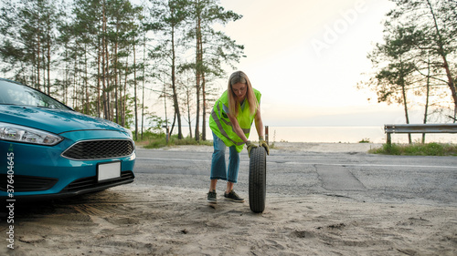 Full length shot of attractive young woman wearing reflective vest, rolling spare tire to change the flat one on the road side