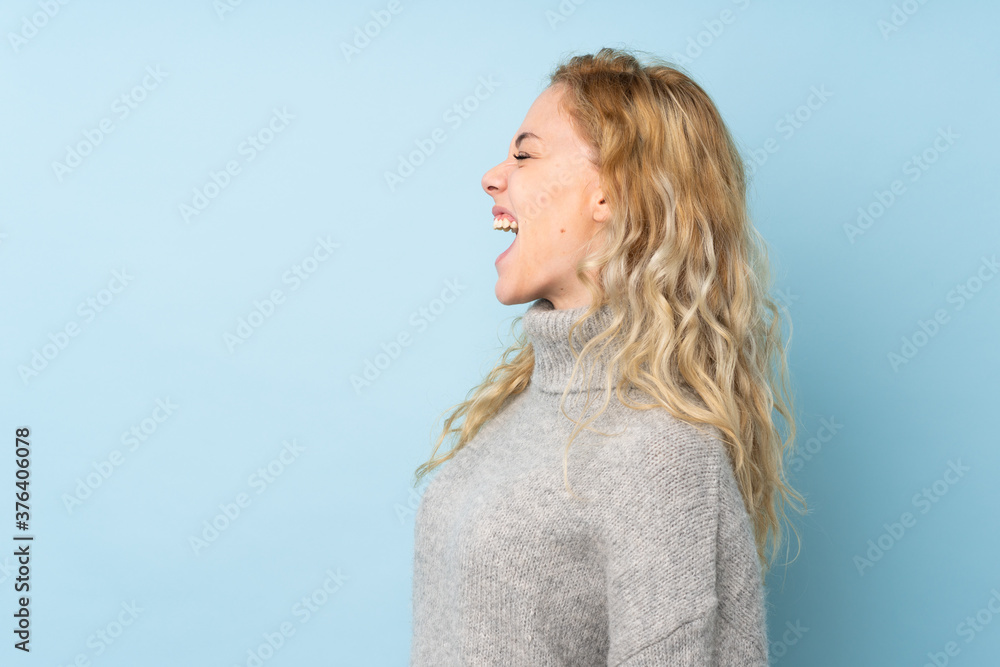 Young blonde woman wearing a sweater isolated on blue background laughing in lateral position Young blonde woman wearing a sweater isolated on blue background laughing in lateral position