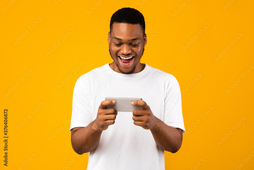 Excited African Guy Using Smartphone Playing Mobile Games, Yellow Background Excited African Guy Using Smartphone Playing Mobile Games, Yellow Background