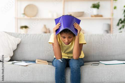 Upset Girl Covering Head With Exercise-Book Sitting On Sofa Indoors photo