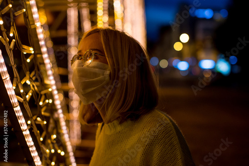 Portrait of a young woman in a yellow sweater with a coronavirus mask, against the background of yellow city lights at night. Bokeh in the background. Woman with sanitizer gel. Coronavirus concept.