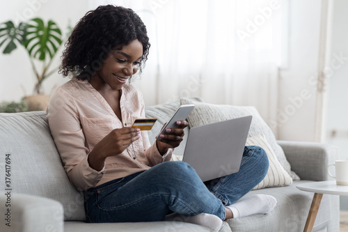 Online Shopping. Cheerful black woman using smartphone, laptop and card at home