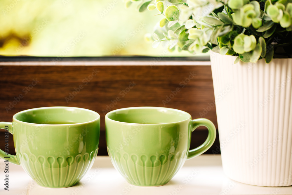 Two green cups with tea stand on a white windowsill next to a plant. Two green cups with tea stand on a white windowsill next to a plant.