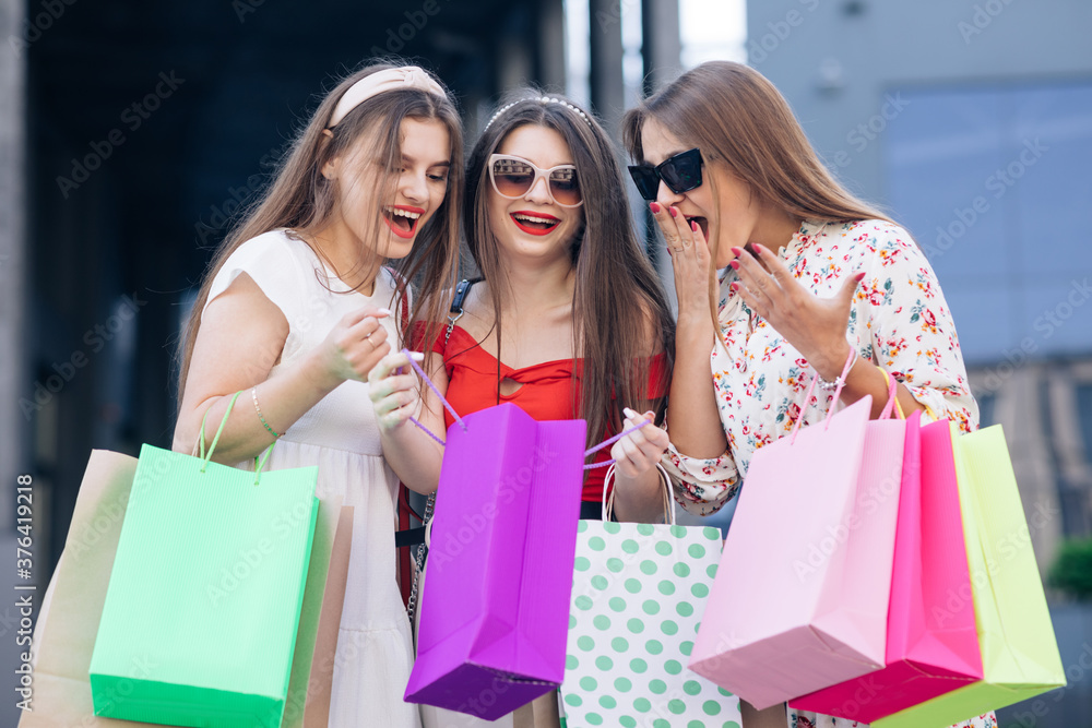 Successful shopping. Casual makeup. A group of young happy cute woman in casual dresses, top and pants walking from the building with yellow, green, purple and pink bags in their hands Successful shopping. Casual makeup. A group of young happy cute woman in casual dresses, top and pants walking from the building with yellow, green, purple and pink bags in their hands