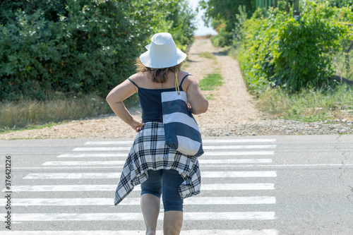 Full woman in a hat and glasses crosses the road at a pedestrian crossing in sunny weather. A female pedestrian with a striped shoulder bag crosses the road. © Elena