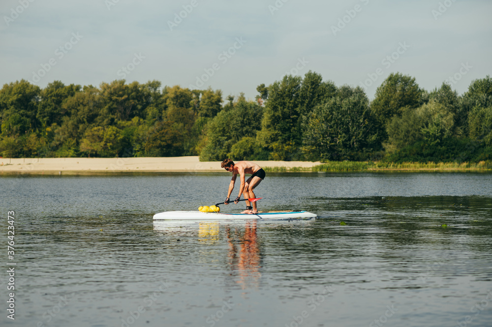 Athletic young man gets on a sup board on the river on a beach background with a tree. Active recreation on the water.