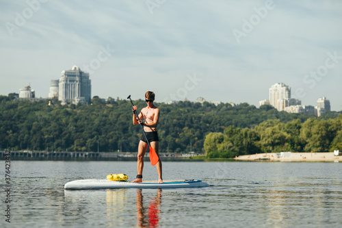 Athletic young man with a naked torso floats on the river on a sup board, rowing with an oar on the background of a beautiful landscape. Active recreation on the water.