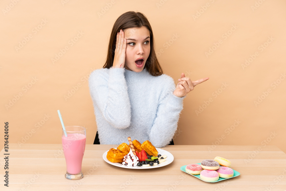 Teenager girl eating waffles isolated on beige background surprised and pointing finger to the side Teenager girl eating waffles isolated on beige background surprised and pointing finger to the side
