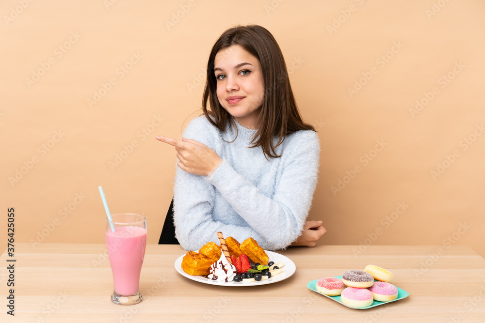 Teenager girl eating waffles isolated on beige background pointing to the side to present a product Teenager girl eating waffles isolated on beige background pointing to the side to present a product