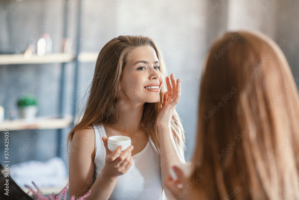 Skin care routine. Lovely blonde girl applying facial cream near mirror at bathroom Skin care routine. Lovely blonde girl applying facial cream near mirror at bathroom