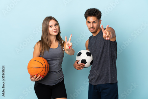 Young couple playing football and basketball isolated on blue background smiling and showing victory sign