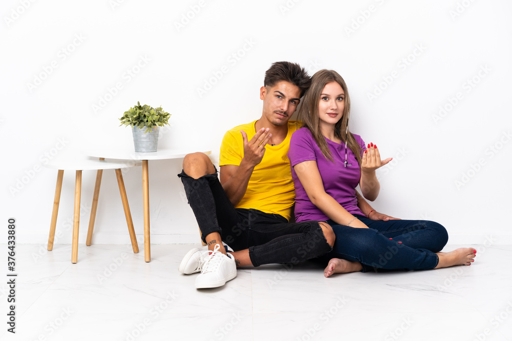 Young couple sitting on the floor isolated on white background inviting to come with hand. Happy that you came Young couple sitting on the floor isolated on white background inviting to come with hand. Happy that you came