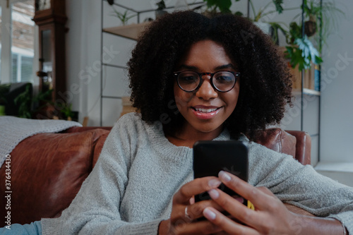Mixed race woman wearing glasses smiling scrolling on smartphone sitting on couch in minimalistic lounge.