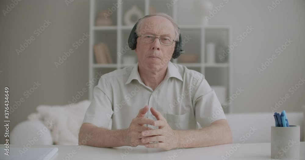elderly male teacher with headphones is speaking during online education by internet, medium portrait of aged speaker elderly male teacher with headphones is speaking during online education by internet, medium portrait of aged speaker