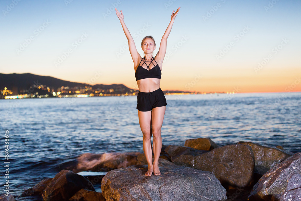 Young woman is practicing stretching on a rock near sea at dawn