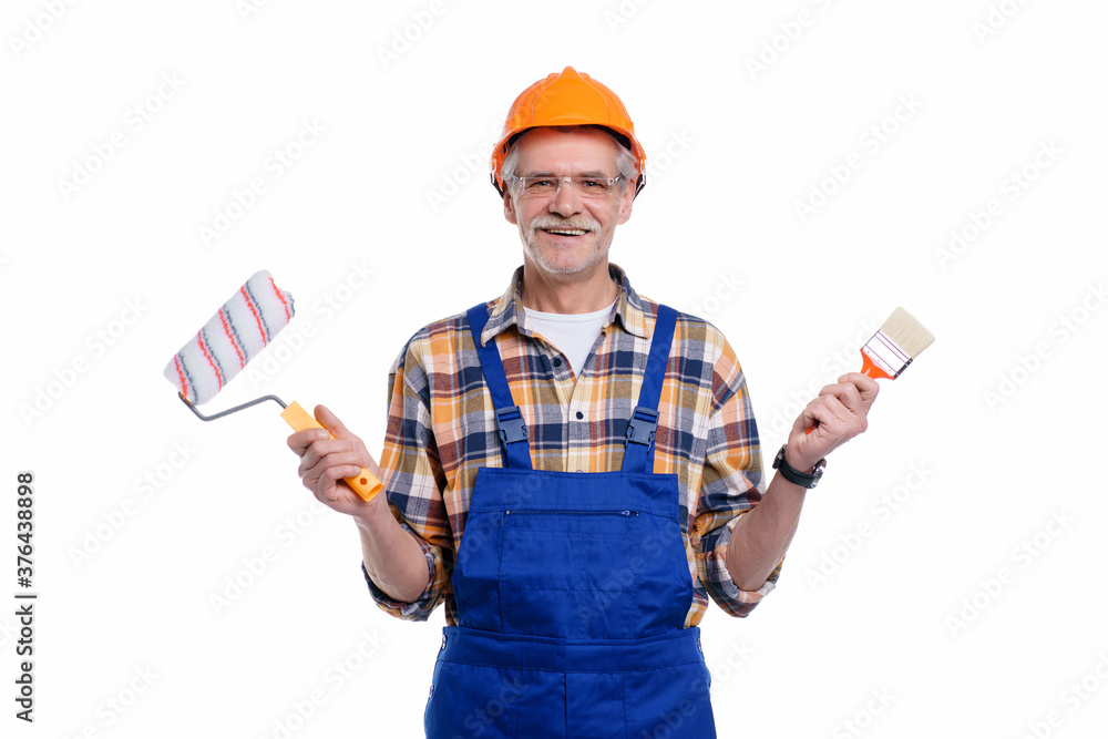 Photo of positive senior male builder holding paint roller and brush in hand, wearing special uniform, apron and protective helmet, isolated on white background Photo of positive senior male builder holding paint roller and brush in hand, wearing special uniform, apron and protective helmet, isolated on white background