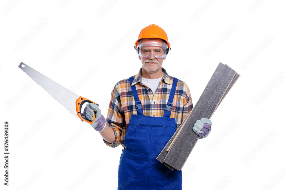 Senior builder in a helmet holding wood boards and saw, looking at camera over white wall background, repair, construction, building, people and maintenance concept Senior builder in a helmet holding wood boards and saw, looking at camera over white wall background, repair, construction, building, people and maintenance concept