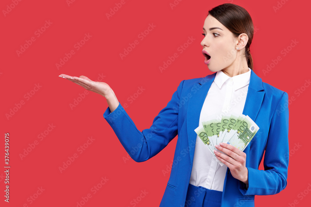 Surprised business woman holding money and showing product, caucasian female wearing blue suit with cash, smiling girl touch euro banknotes on red isolated background Surprised business woman holding money and showing product, caucasian female wearing blue suit with cash, smiling girl touch euro banknotes on red isolated background
