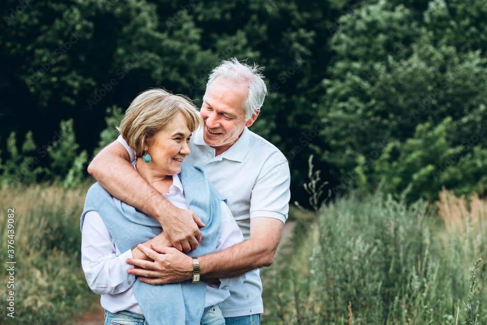 mature couple is walking with dog in park. Elderly couple resting in nature with a dog. Full-length portrait of an elderly man and woman in white shirts and jeans. Stylish and modern grandparents.