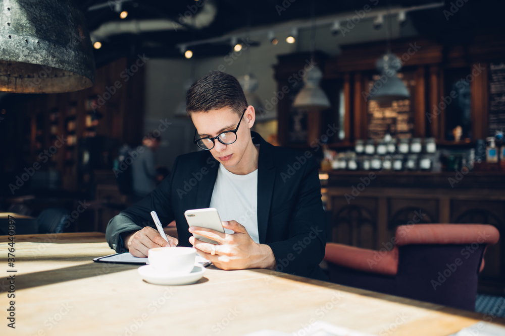 Pondering student with smartphone in hand studying textbook while sitting at cafe