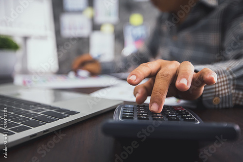 Close up hand of business man press on calculator, accounting and checking on financial graph paper and laptop to calculate profit and budget and income, accountant and counting concept