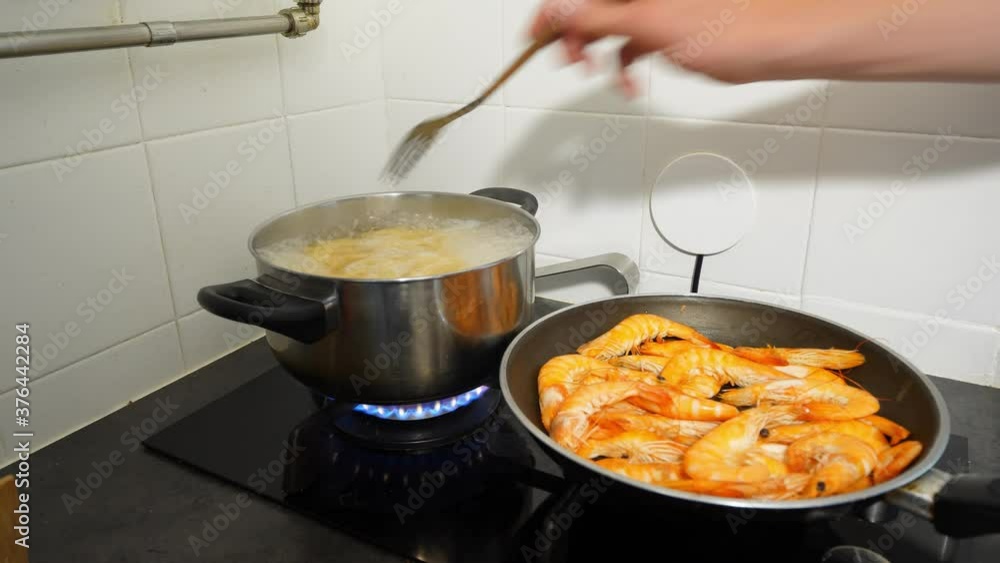 Pasta boil in small pan, shrimps warm up in frying pan. Man prepare food, stir spaghetti with fork, check if it ready. Close up shot of small gas cooktop at apartment house. Pasta boil in small pan, shrimps warm up in frying pan. Man prepare food, stir spaghetti with fork, check if it ready. Close up shot of small gas cooktop at apartment house.