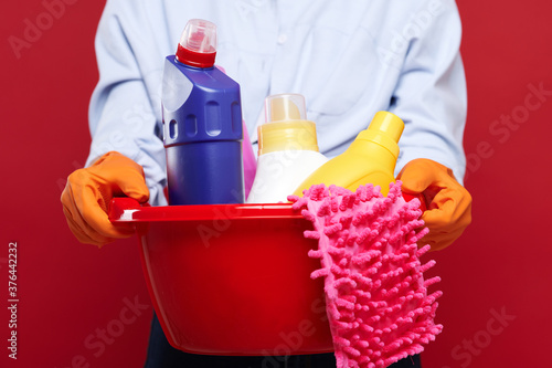Tired housewife wearing casual clothes and latex gloves holding plastic basin with many cleaning products, isolated background © khmelev