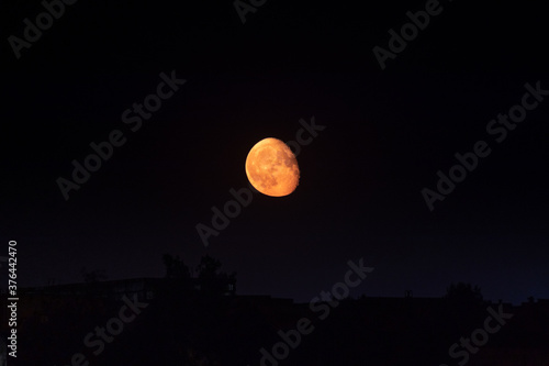 Bright orange moon in waning gibbous phase rising up on dark clear night sky above dark city silhouette. Mystic nighttime black sky with large moon, dark tranquility