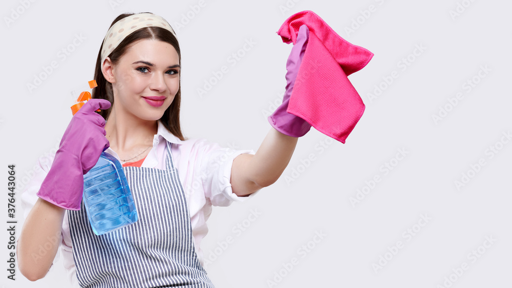 Young smiling housewife in apron and rubber gloves wipes the surface with red rag and using cleaning agent, isolated on white background, concept of cleaning and cleaning companies Young smiling housewife in apron and rubber gloves wipes the surface with red rag and using cleaning agent, isolated on white background, concept of cleaning and cleaning companies