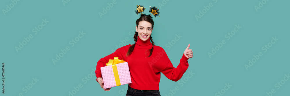 Smiling caucasian girl in red sweater holding christmas gift and showing thumb up gesture in green isolated background Smiling caucasian girl in red sweater holding christmas gift and showing thumb up gesture in green isolated background
