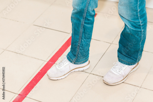 Person stand in red line, legs close-up. Attention line on the floor of the store to maintain social distance. Concept of the coronavirus pandemic and prevention measures
