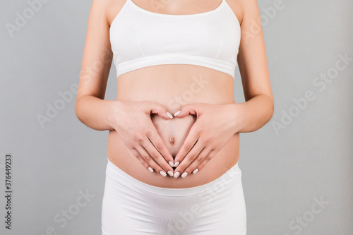 Cropped image of pregnant woman holding her hands in a heart shape on her baby bump at gray background. Motherhood concept. Copy space