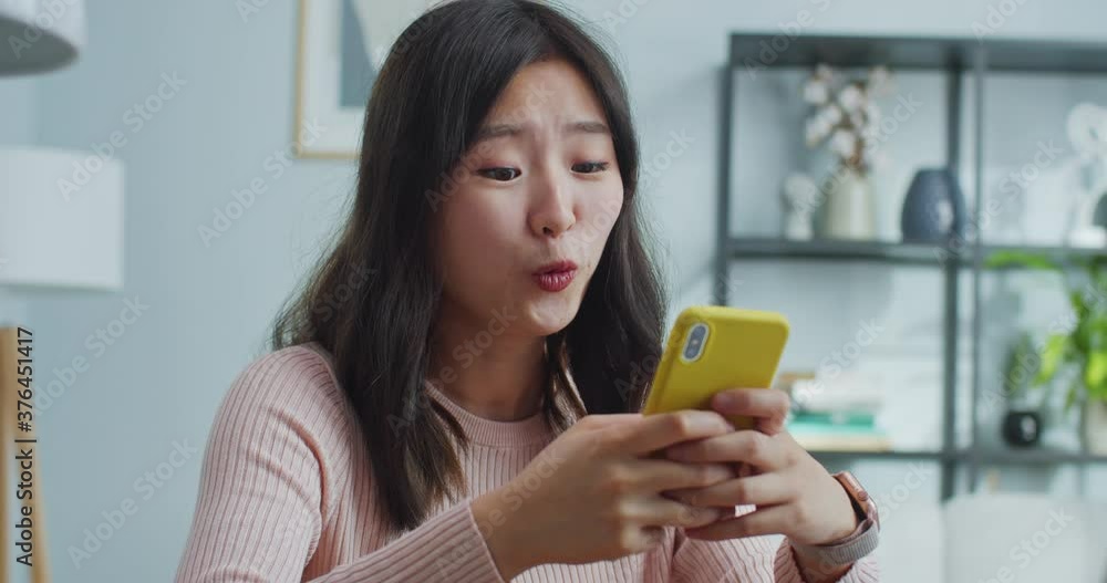 Young Asian woman sitting at table in modern room with minimalist design. Charming girl using smartphone, scroll news feed, chat with friends and watch photos and videos. Technologies and social media Young Asian woman sitting at table in modern room with minimalist design. Charming girl using smartphone, scroll news feed, chat with friends and watch photos and videos. Technologies and social media