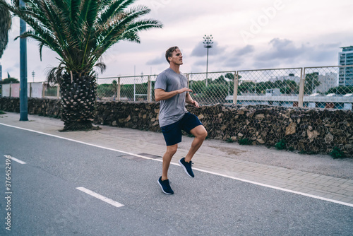 Strong caucasian male athlete leaping on cardio workout slimming and feeling energy for reaching goals on street, sportsman jumping training legs and lower body for running and toning in morning