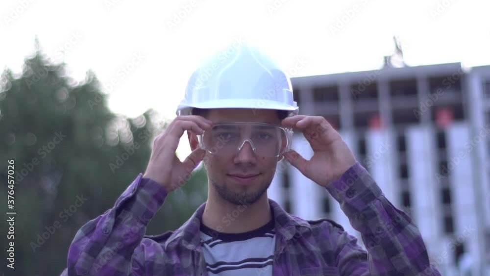 Young bearded caucasian Architect in Hard Hat on the background of the building. Worker who takes off his goggles and Smiling. Young bearded caucasian Architect in Hard Hat on the background of the building. Worker who takes off his goggles and Smiling.