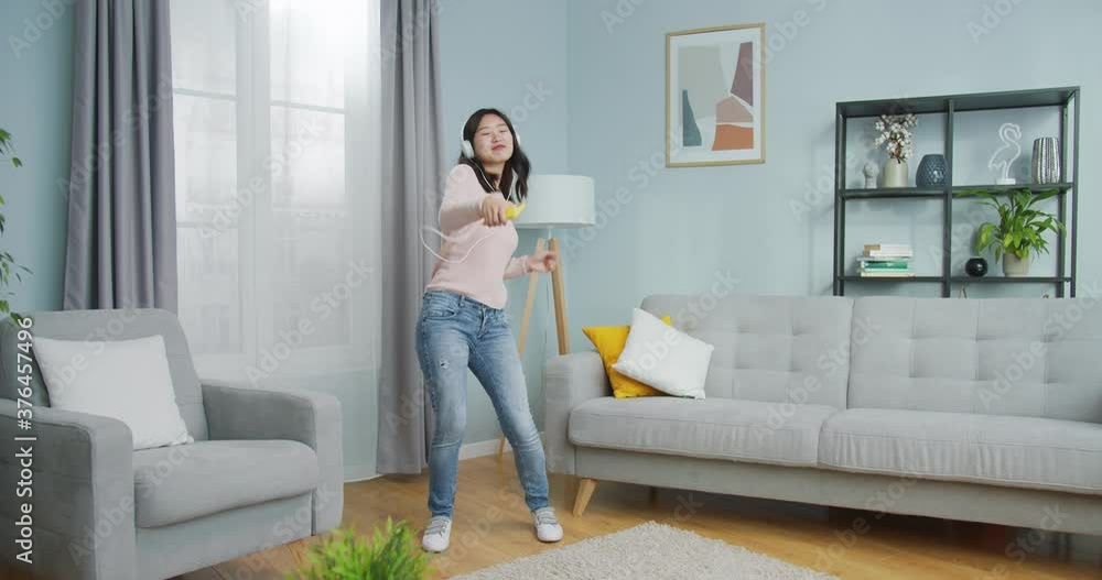 Cheerful Asian girl in headphones having fun in living room with modern minimalist interior at home. Pretty carefree young woman listening to music on smartphone, dancing alone. Hit song, playing app. Cheerful Asian girl in headphones having fun in living room with modern minimalist interior at home. Pretty carefree young woman listening to music on smartphone, dancing alone. Hit song, playing app.