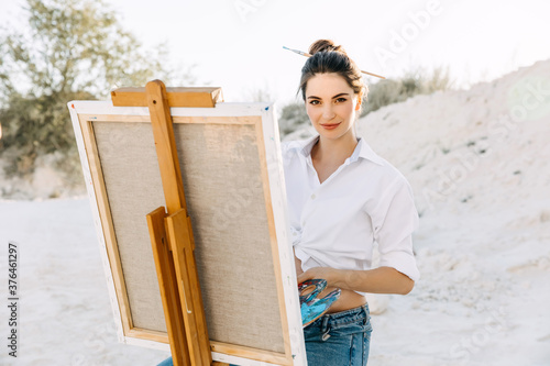 Young woman artist painting on canvas on a easel, holding a palette.