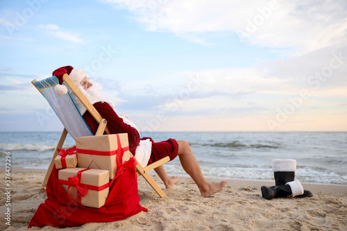 Santa Claus with bag of presents relaxing in chair on beach, space for text. Christmas vacation
