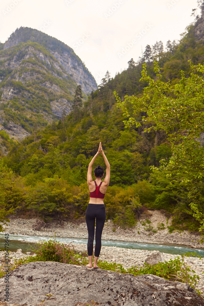 Naklejka premium Yoga classes in nature. The concept of playing sports alone. Social exclusion. A woman does yoga on rocks, near a mountain river flows