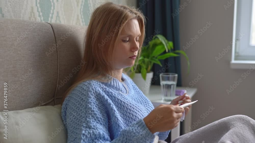 Sick young blonde woman lies in bed at home under blanket and check thermometer. Headache, flu, cold, virus. Sick young blonde woman lies in bed at home under blanket and check thermometer. Headache, flu, cold, virus.
