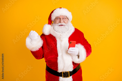 Photo of excited grey white beard hair santa claus use smartphone raise fists celebrate x-mas christmas jolly holly lottery victory wear cap headwear isolated bright shine color background