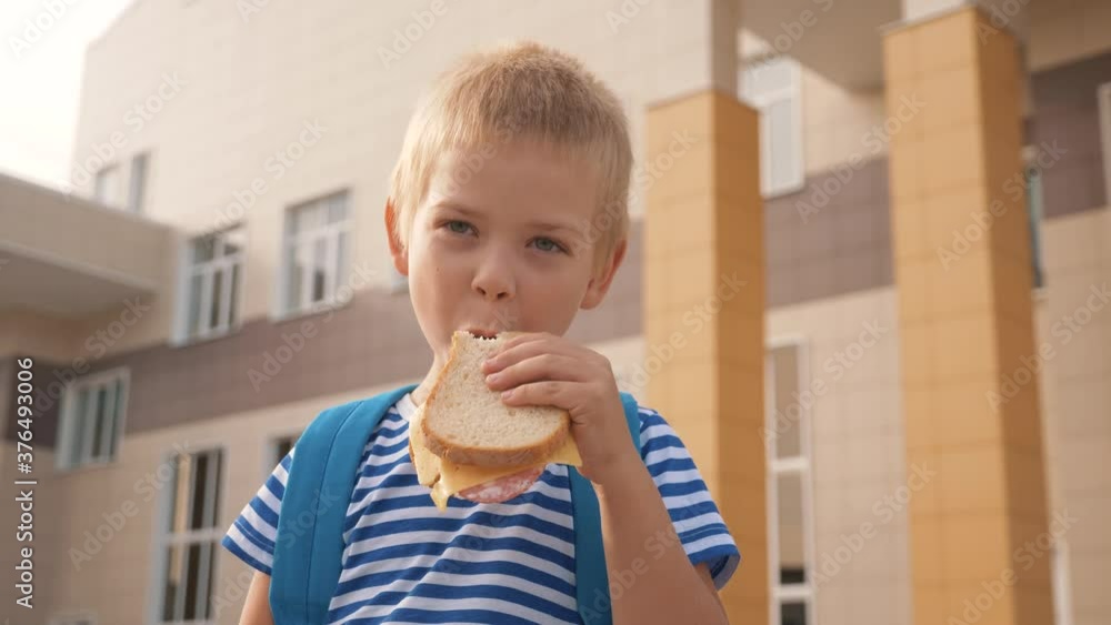 Happy kid eating a sandwich outdoors at school. Healthy school breakfast for the child. Hungry kid eating outside on the background of the school. Street fast food. Food for lunch. A delicious burger Happy kid eating a sandwich outdoors at school. Healthy school breakfast for the child. Hungry kid eating outside on the background of the school. Street fast food. Food for lunch. A delicious burger