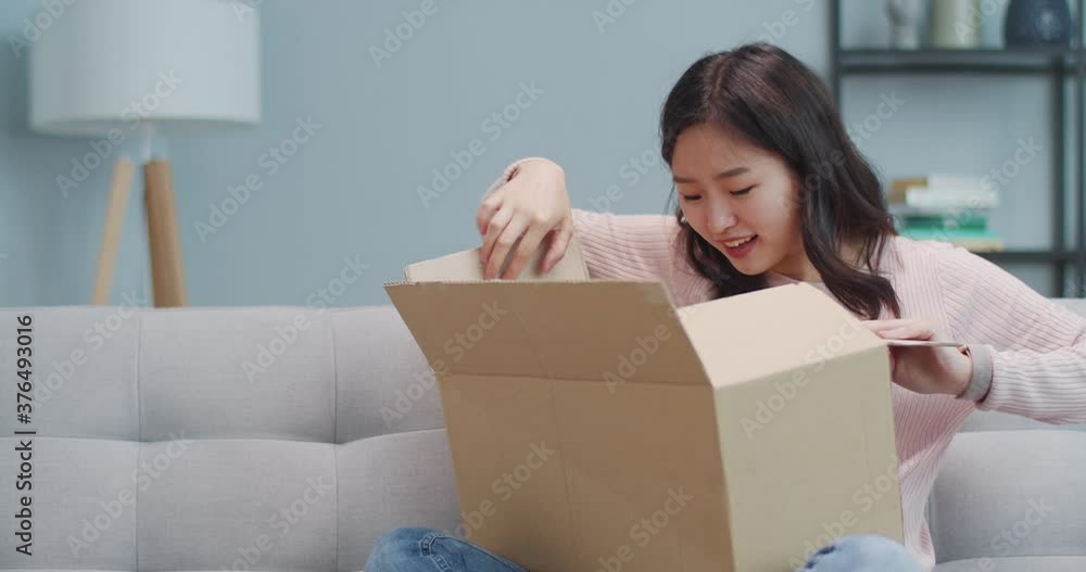 Happy amazed young Asian girl sitting on couch and opening cardboard box at home. Excited surprised woman customer receiving parcel, order. Courier, delivery, post, e-commerce. Happy amazed young Asian girl sitting on couch and opening cardboard box at home. Excited surprised woman customer receiving parcel, order. Courier, delivery, post, e-commerce.