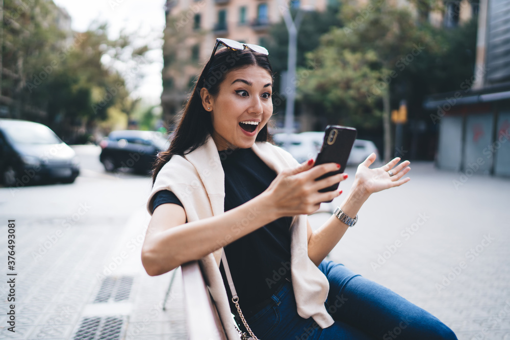 Amazed emotional asian hipster girl surprised with sales in web store browse on smartphone outdoors, smiling overjoyed blogger happy about number of followers checking notification on cellular