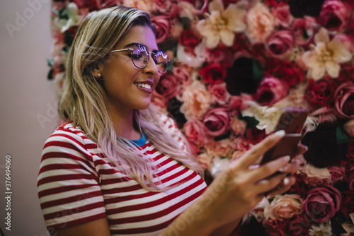 Pretty Spanish female blogger in optical glasses with neon lights reflection using modern cellphone gadget for writing sms message, millennial girl browsing website for making online booking