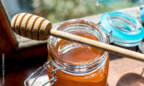 A wooden honey spoon lies on an open glass jar of floral sticky orange honey on a wooden windowsill
