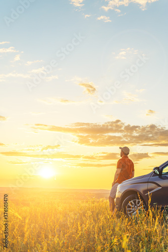 A young man in a shirt enjoys the sunset. A man and his car in a field against the backdrop of a brightly lit sunset sky. Local travel concept. Vertical orientation, space for text.
