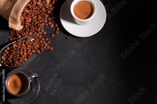 Two espresso cups, coffee beans, metal scoop, burlap bag on black table background with copy space. Arabica grains, top view. Coffee shop, caffeine, hot drink concept